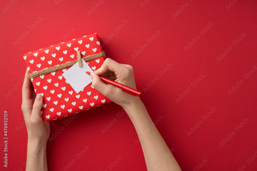 Obraz premium First person top view photo of valentine's day decor female hands writing love letter attached to giftbox in red wrapping paper with heart pattern and twine on isolated red background with empty space