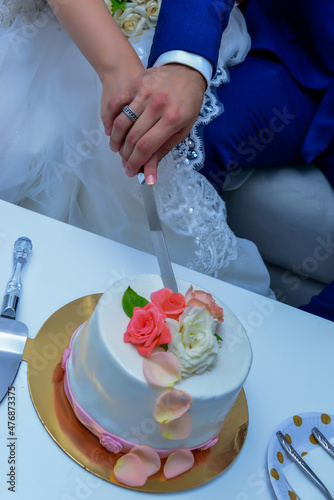 bride and groom cut the wedding cake. The cake is decorated with beige and peach-colored roses. groom is dressed in blue wedding suit and bride in a white wedding dress..