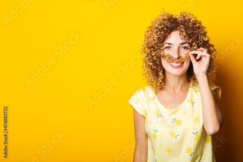 cheerful curly young woman doing mustache with hair on yellow background.