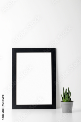 A mock-up of a picture frame with a black wooden frame on a white table, a small succulent. Show text or product. © Татьяна Кочкина