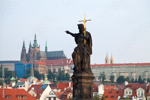 Stone statue on Charles bridge pointing to the St.Vitus Cathedral over red house roofs. Czeck Republick, Prague.