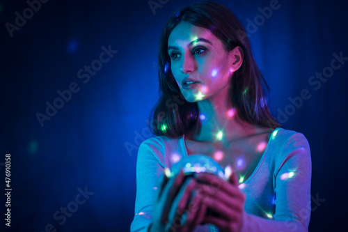A young brunette Caucasian woman in a white t-shirt on a black background. Neon lights, illuminated with a disco light in hand, sweet look