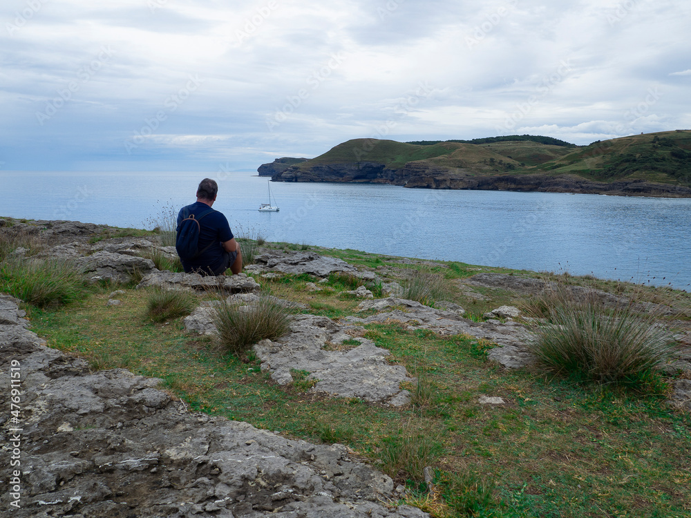 Vistas de una persona sentada de espaldas mirando un barco en el mar azul y la costa en la zona de la Cueva de la Ojerada en Cantabria, España, verano de 2020
