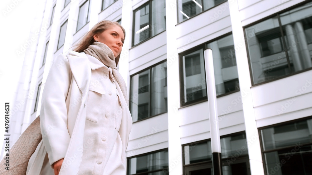 A girl in a white coat and a scarf is walking down the street, smiling. Business center in the background. Success