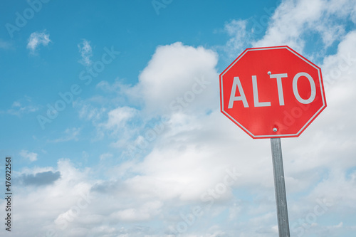 Red Stop sign in Spanish right aligned against the blue cloudy sky background. Alto, wallpaper for text.