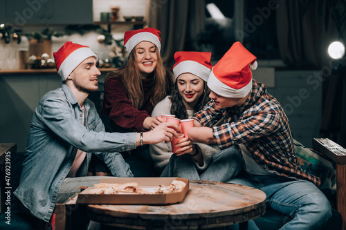 A young man with a disability with amputated arms celebrates the New Year with friends. Young people sit on the couch in the living room with pizza, clink glasses