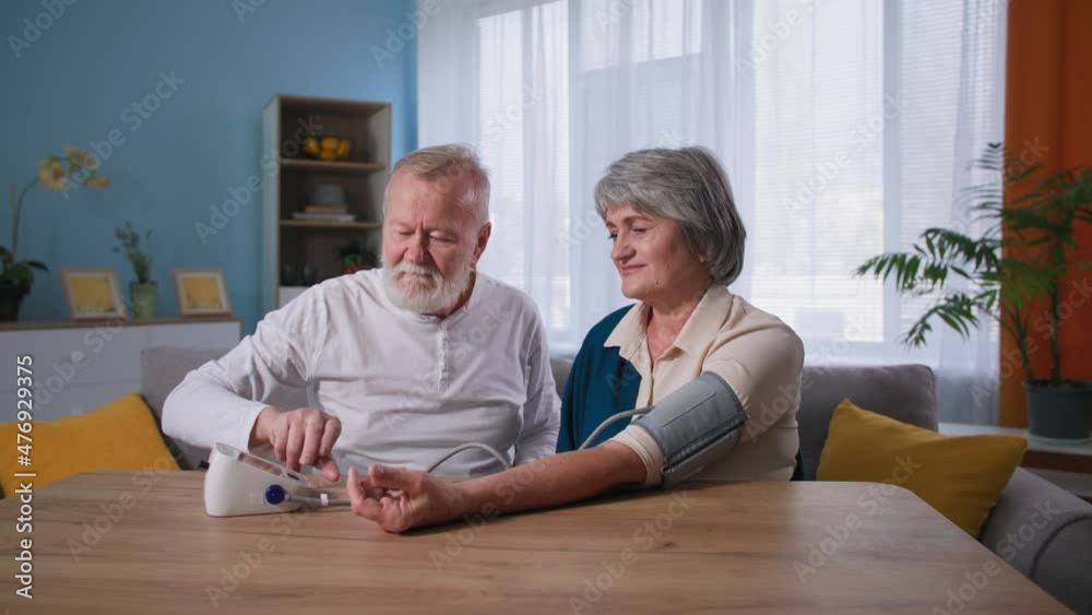 elderly caring man checks blood pressure of his old wife with hypertension using a tonometer at table, old couple take care of health at home elderly caring man checks blood pressure of his old wife with hypertension using a tonometer at table, old couple take care of health at home