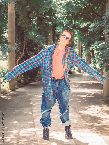 A girl with two braids in a plaid shirt with overly long sleeves and huge jeans stands spreading her arms to the side in the park on a green alley. Post-processing. Artistic effect photo