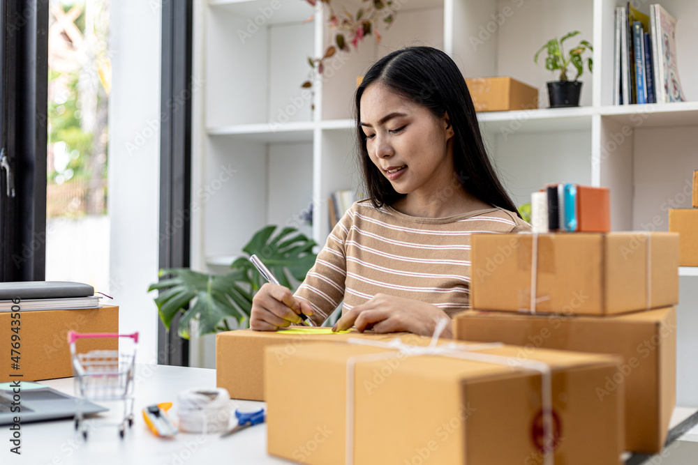 Asian woman writing customer's shipping information on parcel box, she owns an online store, she ships products to customers through a private courier company. Online selling concept. Asian woman writing customer's shipping information on parcel box, she owns an online store, she ships products to customers through a private courier company. Online selling concept.