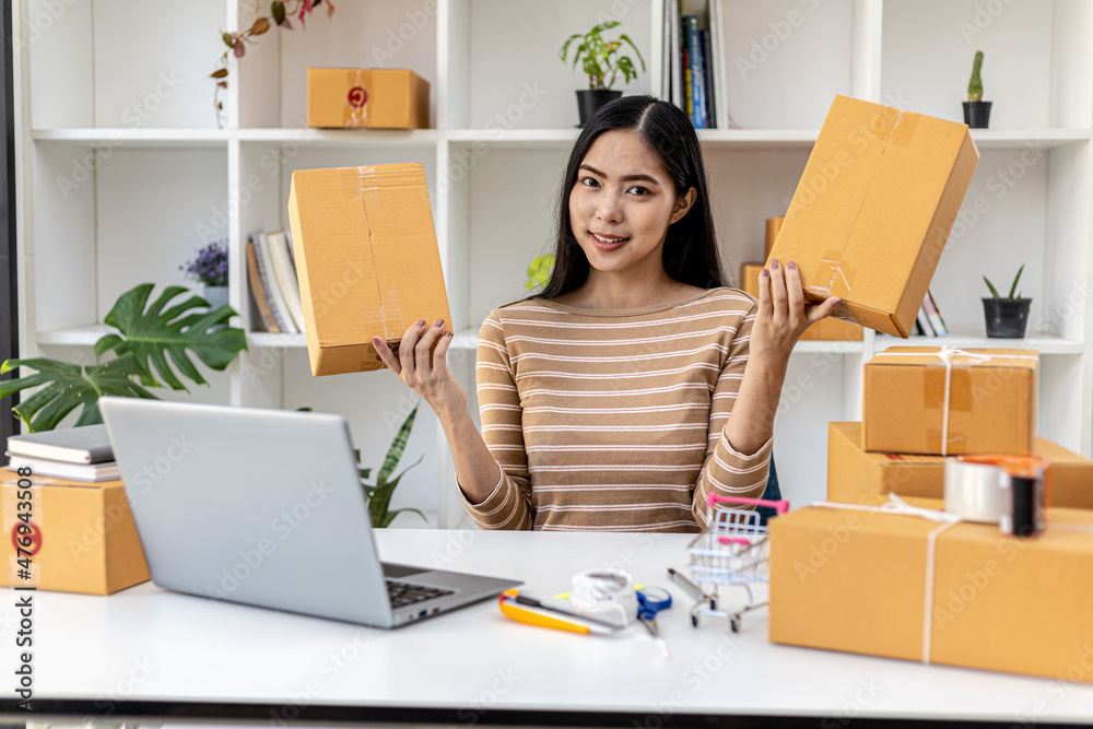 Portrait woman holding two parcels on a table with a laptop and a bunch of parcels, she's an online seller, she's packing up a customer's order. Concept of selling products online and packing products Portrait woman holding two parcels on a table with a laptop and a bunch of parcels, she's an online seller, she's packing up a customer's order. Concept of selling products online and packing products