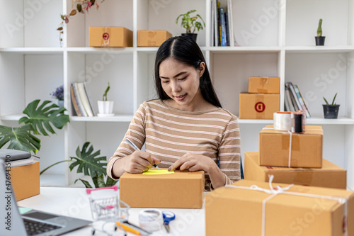 Asian woman writing customer's shipping information on parcel box, she owns an online store, she ships products to customers through a private courier company. Online selling concept.