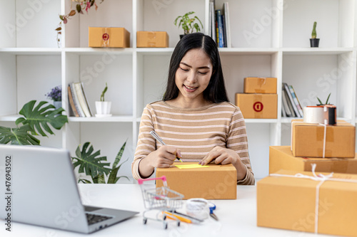 Asian woman writing customer's shipping information on parcel box, she owns an online store, she ships products to customers through a private courier company. Online selling concept.