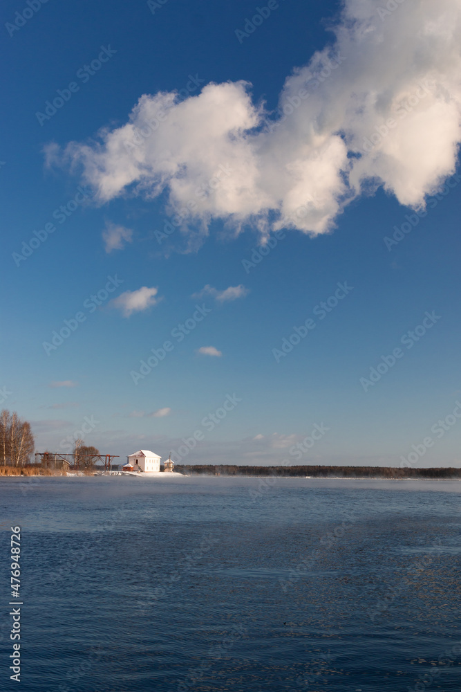 Winter photo of a small white fishing house with several trees.. There is snow on the ground, and there is some ice on the lake. bright blue sky with clouds.