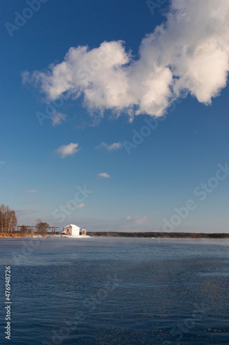 Winter photo of a small white fishing house with several trees.. There is snow on the ground, and there is some ice on the lake. bright blue sky with clouds. © Svetlana