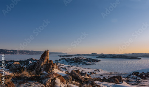Early winter morning in Siberia. The sky is highlighted in orange. Sun glare on the ice of a frozen lake. Camping on a snowy shore. In the foreground are picturesque granite rocks, dry grass. Baikal. 