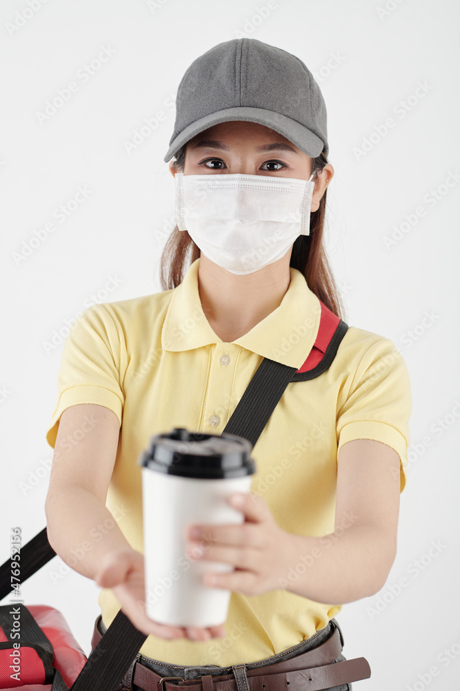 Portrait of female courier in medical mask giving cup of coffee in disposable cup