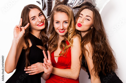 Three best friends celebrate birthday indoor wearing elegant evening dress and have bright make up. Girls hugging and Showing signs with their hands. Looking at camera and smiling. Inside.