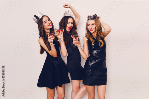 Three beautiful elegant women celebrate hen-party and drinking cocktails. Best friends wearing black  stylish evening dress,  high heel shoes ,crown on head .Bright make up, red lips. Inside.