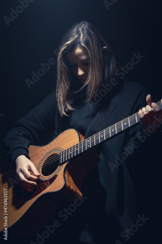 A young woman with an acoustic guitar in the dark under a ray of light.