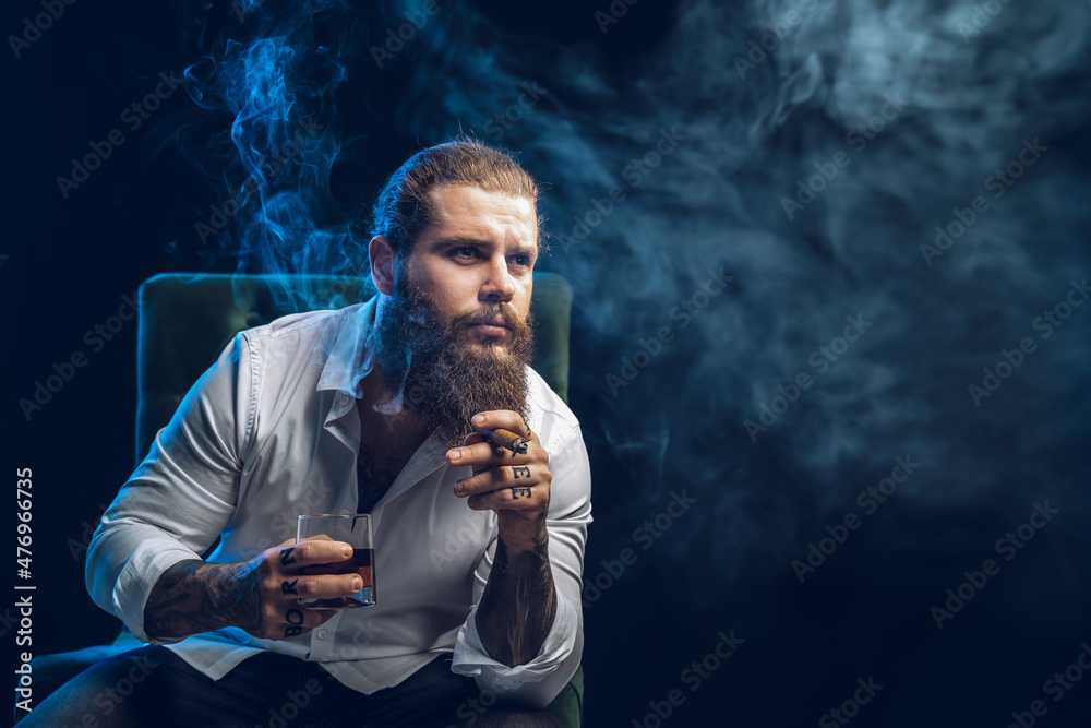 Brutal hipster bearded man smoke a cigar drinks whiskey, dressed in white shirt. Studio shot