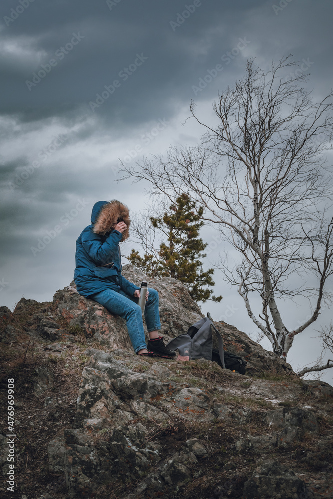 Young woman in a blue down jacket drinks tea from a thermos on a mountain top under a gloomy gray sky