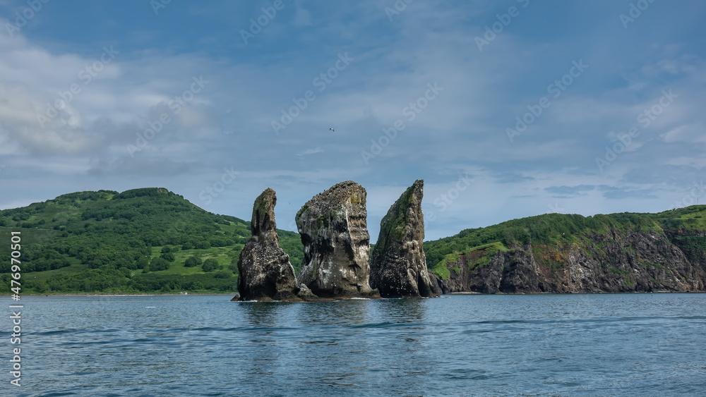 High picturesque cliffs rise above the ocean. Bird nests on the rocks. The hilly coast of Kamchatka against the background of blue sky and clouds. Three Brothers.