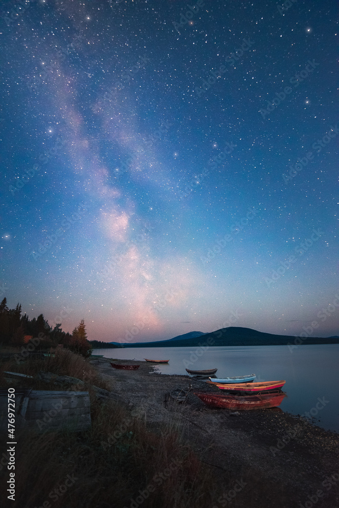 Milky Way and sky full of stars above the lake and moored boats with mountains on the horizon