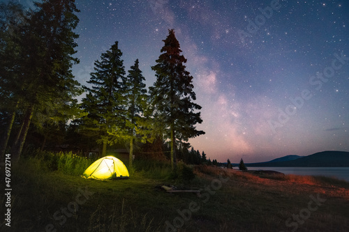 Milky Way and sky full of stars above the luminous tent under the trees by the lake