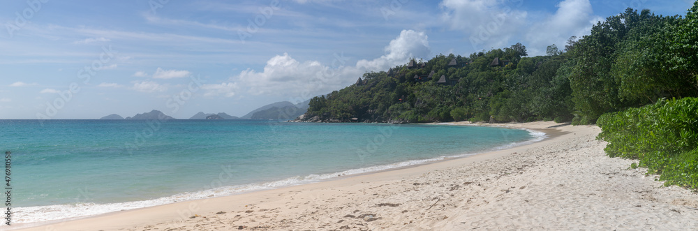 Large-format panoramic photo with high resolution of a typical Seychelles landscape. A perfect snow-white beach. In the distance, on the green hills, there are bungalows with thatched roofs.