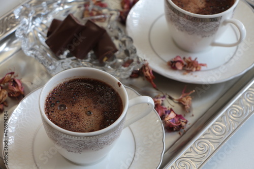 Two white cup of Turkish coffee on silver tray.Traditional Turkish Coffee.Coffee foam.Small porcelain coffee cup with silver detail.Dried pink flowers and chocolates on a glass plate. Selective focus