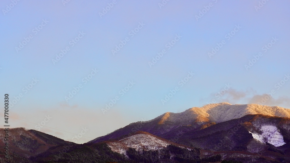 群馬県高山村の雪が積もり始めた山に夕日の光がオレンジ色にうつっている風景