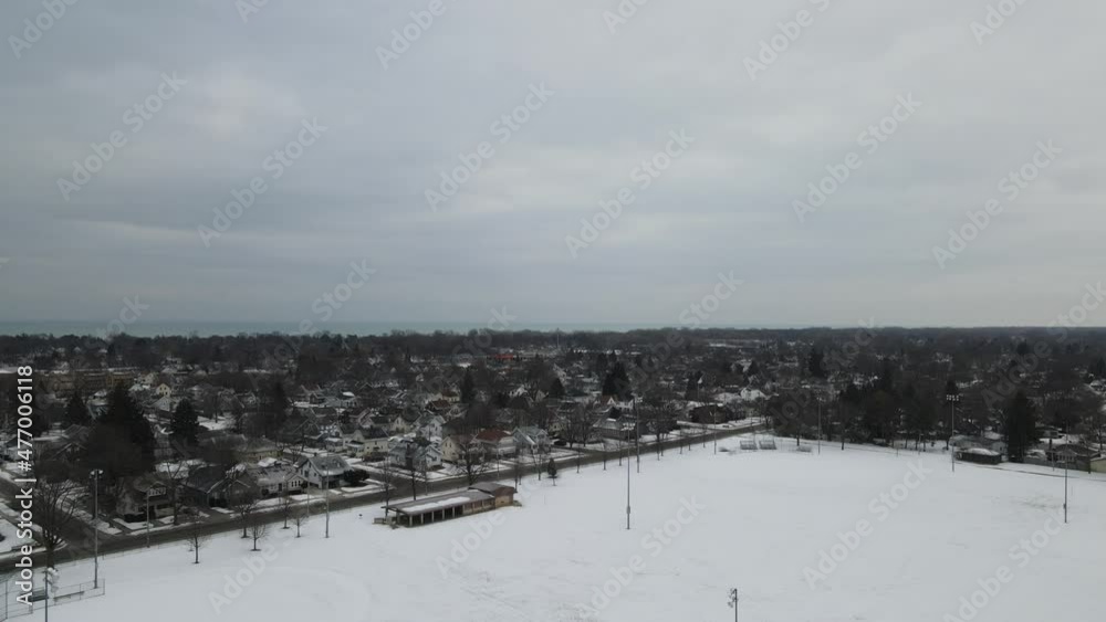 Aerial view over ball field covered in snow. Park shelter empty and waiting. Residential neighborhood surrounding park. Cloudy overcast sky of winter. Aerial view over ball field covered in snow. Park shelter empty and waiting. Residential neighborhood surrounding park. Cloudy overcast sky of winter.