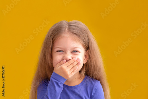 Portrait of laughing caucasian little girl child kid of 5 years looking at camera covering mouth by hand giggle on yellow background photo