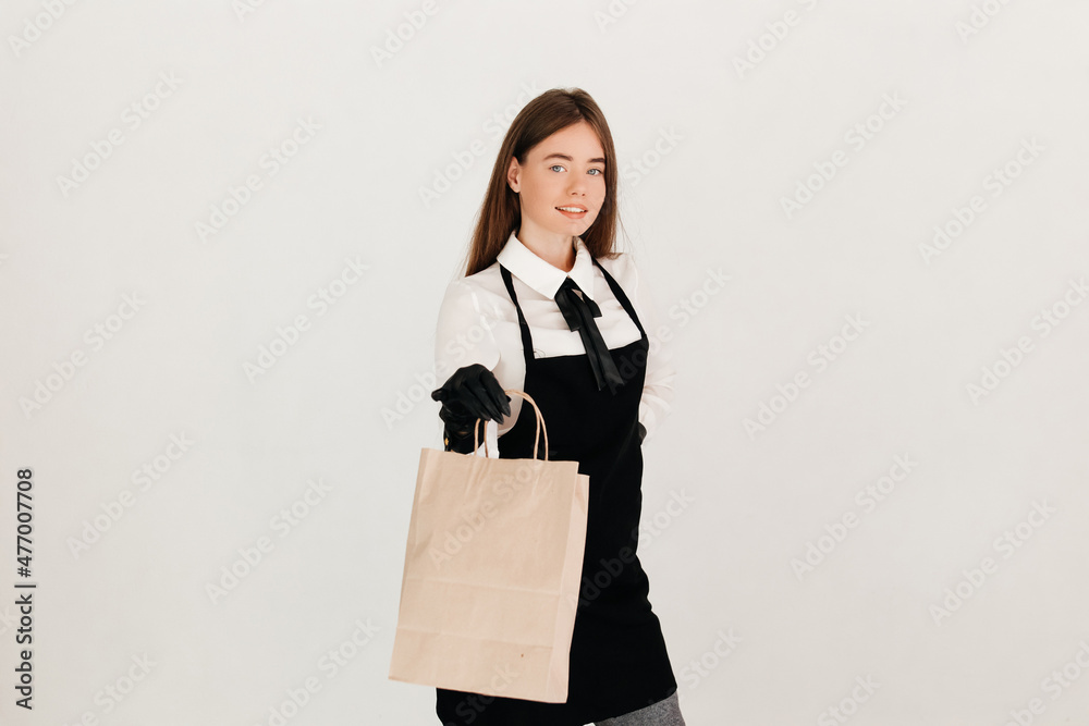 Young attractive waitress in a uniform and protective medical gloves holding a paper craft bag with delivery food, smiling and looking at the camera Young attractive waitress in a uniform and protective medical gloves holding a paper craft bag with delivery food, smiling and looking at the camera