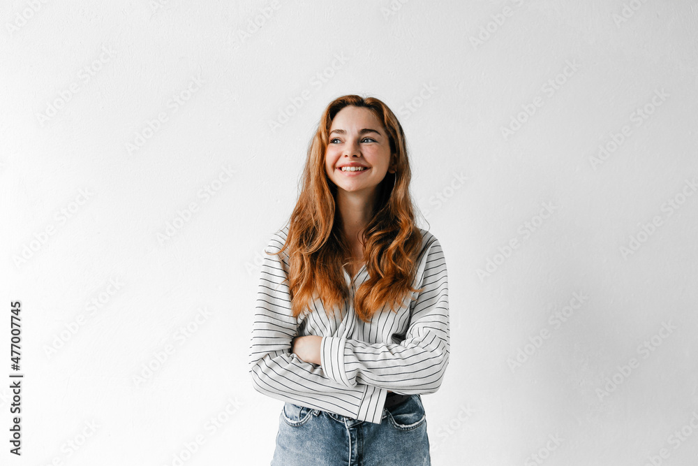 Emotive portrait of smiling redhead woman with curly hair, crossing her arms and looking to the side, standing against white background Emotive portrait of smiling redhead woman with curly hair, crossing her arms and looking to the side, standing against white background