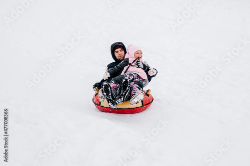  kid smiling and sliding down hill on sled with father looking away wearing warm winter clothes in forest. Astonishing background full of white color and snow. 