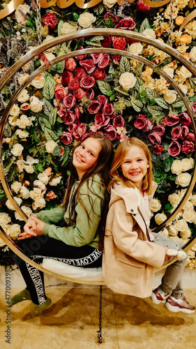 two little girls sit on a swing against a wall of flowers. 
