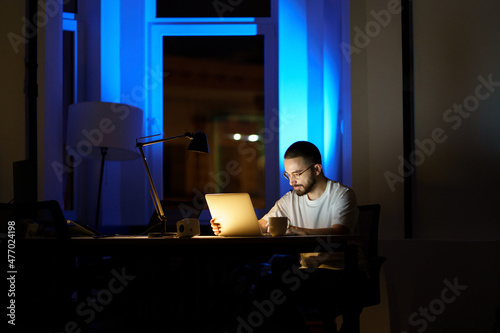 Focused young businessman in eyeglasses work on laptop late at night preparing for new startup project presentation. Concentrated freelance programmer stay overtime to meet application launch deadline