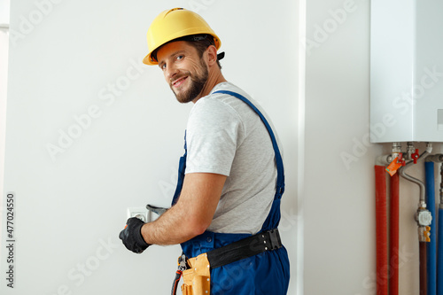 Confident electrician in hard hat and protective gloves smiling at camera while installing electrical socket in a room