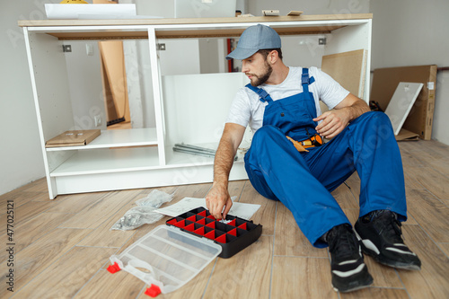 Focused handyman in overalls sitting on the floor while assembling kitchen cabinet in apartment