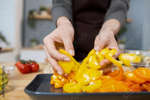 Hands of female putting chopped fresh yellow capsicum on tray while cooking vegetarian stew for dinner in the kitchen