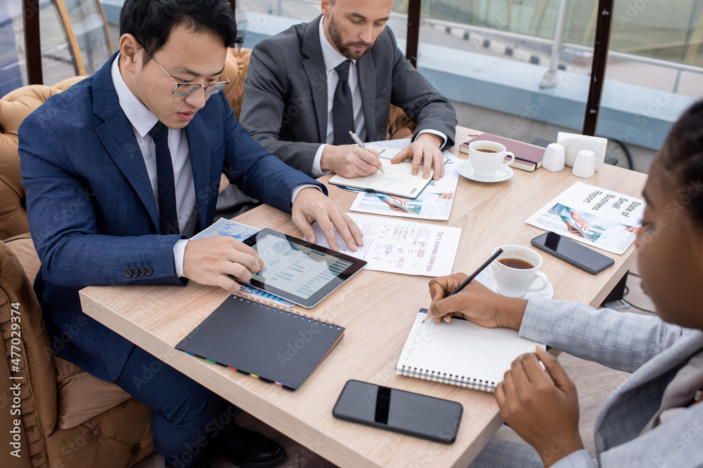 Young Chinese businessman scrolling through financial charts in tablet while sitting by table among intercultural coworkers Young Chinese businessman scrolling through financial charts in tablet while sitting by table among intercultural coworkers