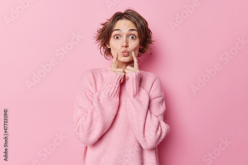 Beautiful woman with bob hairstyle keeps lips rounded index fingers on cheeks has romantic look awaits for kiss wears warm cashmere jumper isolated over pink background. Face expressions concept