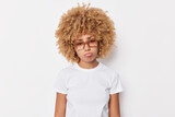 Upset displeased curly haired European woman looks sadly at camera purses lips feels bored and discontent wears casual t shirt and spectacles isolated over white background. Negative emotions