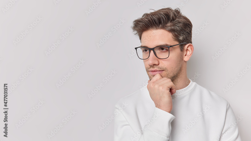 Horizontal shot of thoughtful young man holds chin looks attentively away ponders on something wears spectacles and jumper isolated over white background copy space for your promotional content Horizontal shot of thoughtful young man holds chin looks attentively away ponders on something wears spectacles and jumper isolated over white background copy space for your promotional content