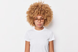 Portrait of serious curly haired young woman looks attentively at camera wears spectacles and basic t shirt poses against white background has self confident expression listens you attentively