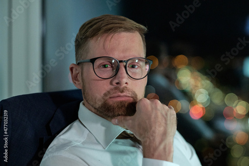 Contemporary bearded businessman in eyeglasses looking at you while sitting by workplace late at night