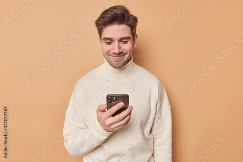 Happy man enjoys messaging with girlfriend via smartphone types sms connected to wireless internet wears turtleneck isolated over beige background. People technology and communication concept © wayhome.studio 
