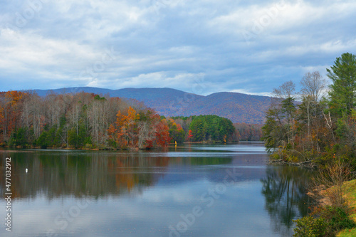 A windsurfer navigates on a pristine mountain lake with colorful clouds and autumnal colors in the trees.