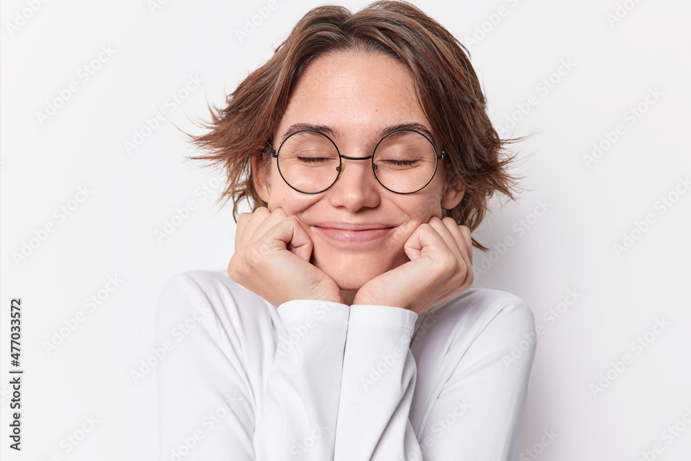 Portrait of charming pleased brunette woman keeps hands under chin closes eyes has dreamy expression recalls something in mind wears rounded spectacles casual jumper isolated over white background